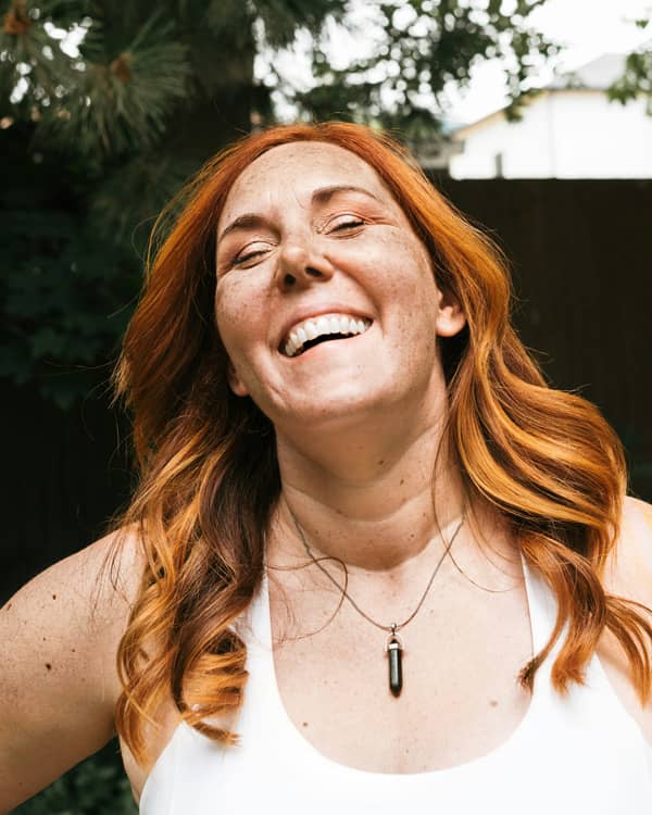 A woman with long red hair and freckles is laughing joyfully outdoors. She is wearing a white tank top and a necklace with a pendant. The background features greenery and a hint of a house, creating a lively and natural setting.